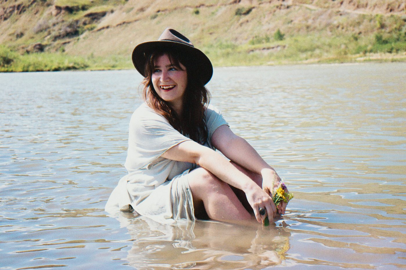 Raine smiling and looking over her shoulder, sitting in shallow river water in a wide-brimmed hat, holding a small bunch of wildflowers, with grassy hills behind her.